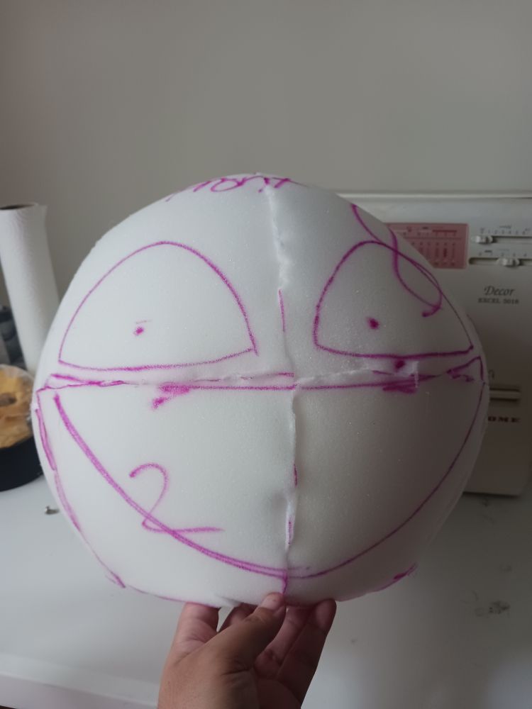 Upholstery foam glued into a round head base with purple marker showing where eyes and a mouth will be with dots in the eyes making the base look unsettling. It is on a white table being held up slightly by the maker's hand
