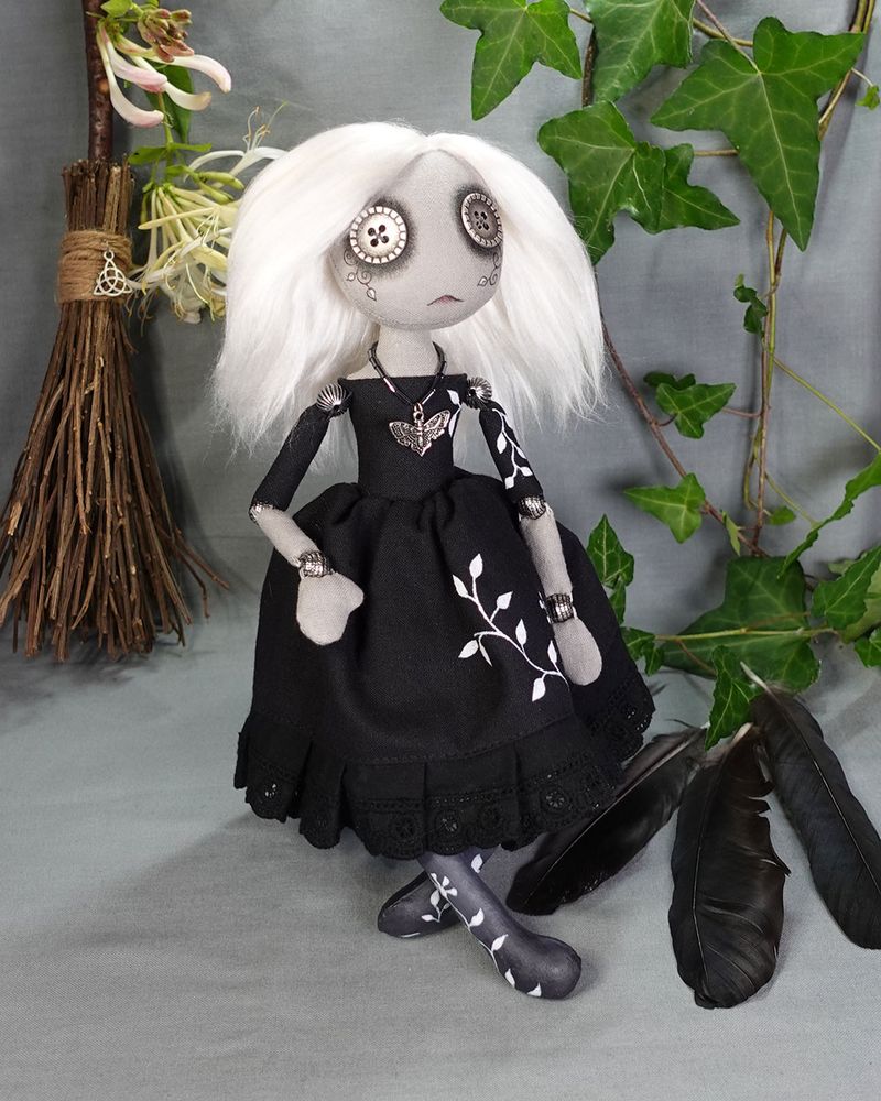 a gothic button eyed textile art doll with white hair, black dress with hand painted vines and moth necklace, on a background of ivy crow feathers and a broom