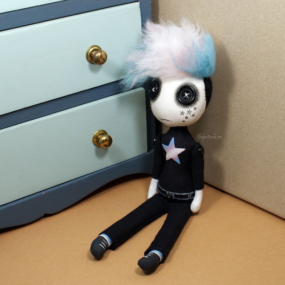 a button eyed art doll with spiky hair in trans flag colours, wearing black shirt and pants with trans flag star on chest