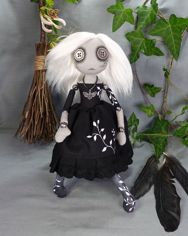 a gothic button eyed textile art doll with white hair and black dress with hand painted vines on a background of ivy crow feathers and a broom