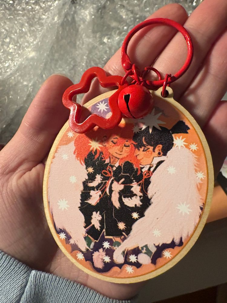 Wooden charm showing aki and angel being tied together by red string, there is a red bell and star embellishment 