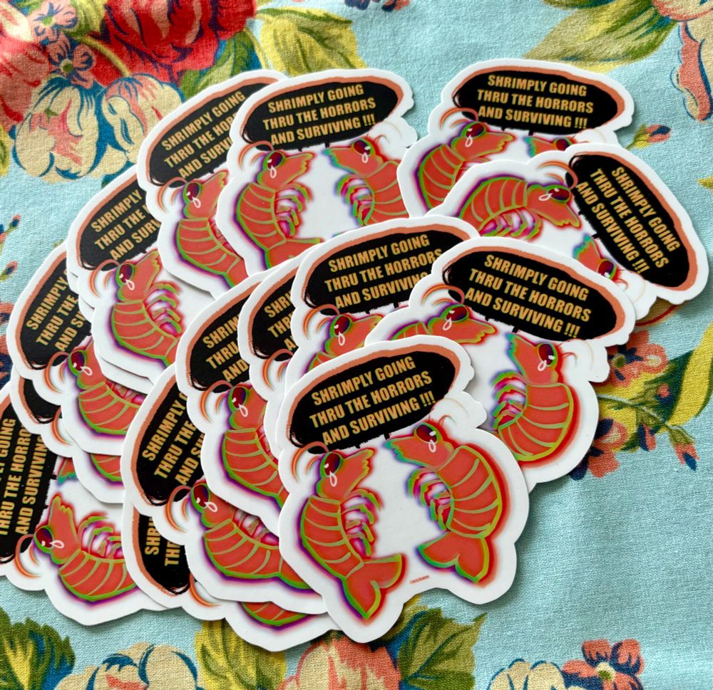 Pile of stickers with two crying shrimp that say “shrimply going thru the horrors and surviving!!!” On a teal floral patterned cloth 