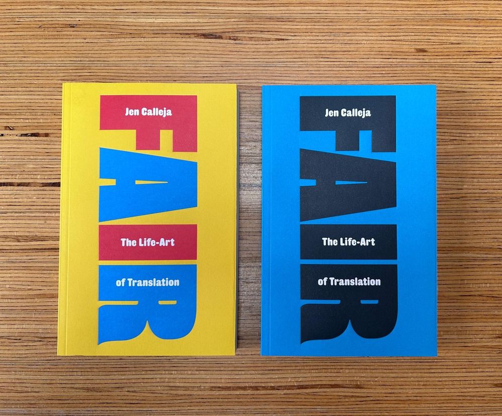 Two books from above on a wooden table. They’re identical but in different colours. The one on the left is yellow with red and green chunky text that says Fair, the one on the right is blue with chunky black text. Fair the life-art of translation by Jen Calleja