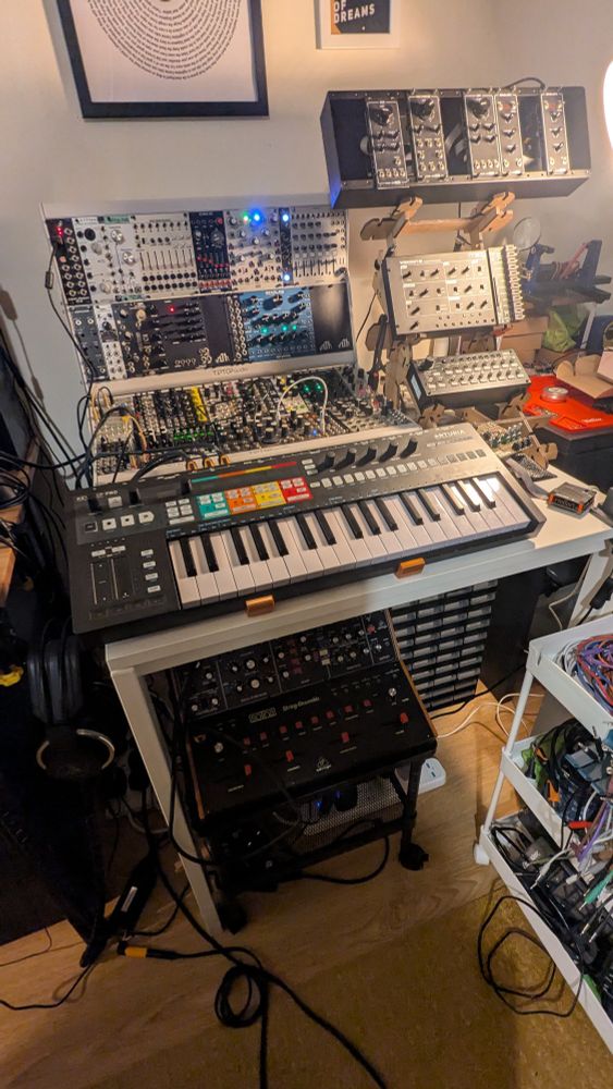 A home studio setup including two 6U 104HP cases full of Eurorack modules; a three-tier wooden stand housing a half-constructed Erica Synths/MKI DIY edu synth, a Moog Werkstatt-01, and a Korg SQ-1 sequencer; a Keystep Pro, a Behringer Solina, and a Behringer Model D.