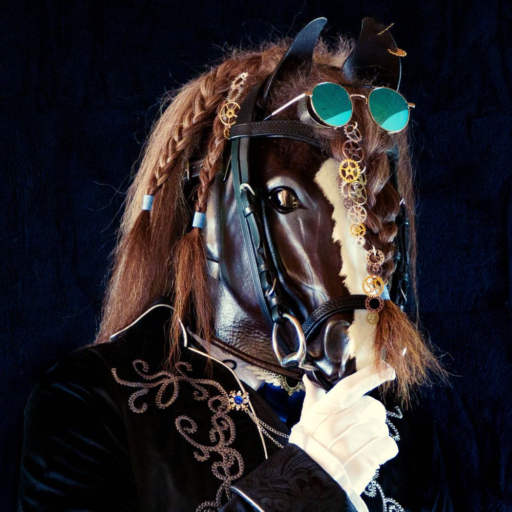 Portrait of someone wearing a horse costume. Head and shoulders visible before black background. Steampunk decoration. Cogwheels in the mane and used as ear-rings. Blue colored sunglasses on head. Wearing a bridle. Hand wearing white gloves near mouth in a thoughtful gesture. Body outfit is a black tailcoat with intricate silver embroidery and blue gemstones with silver chains