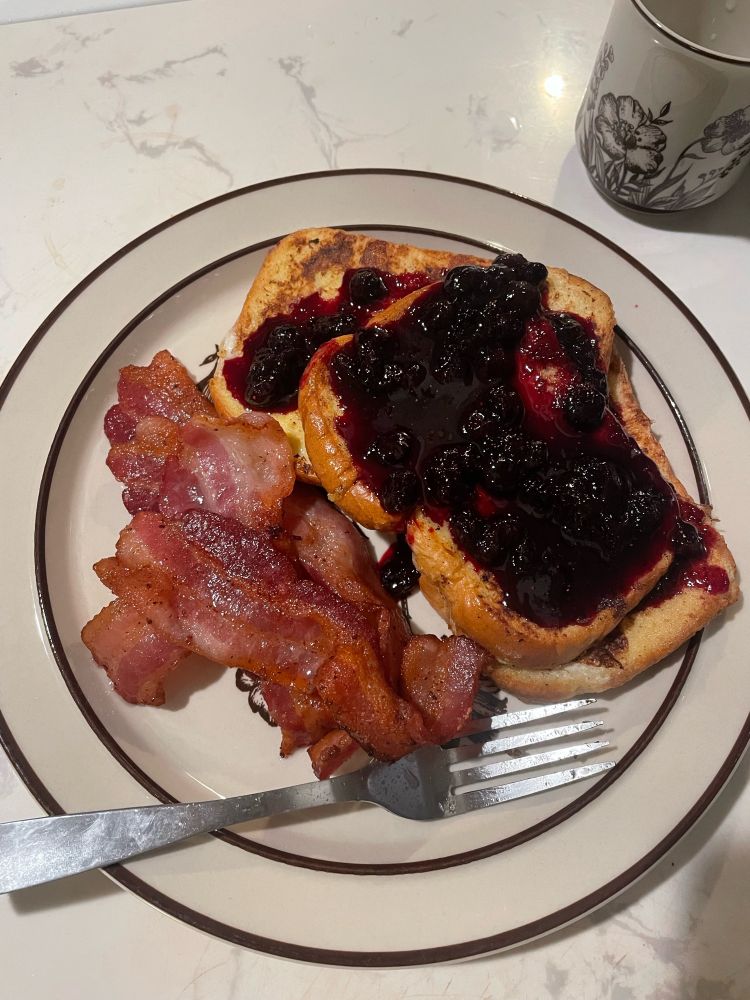 A top down shot of a plate with bacon on one side, and french toast with blueberry compote on the other side. A fork rests against the bottom of the plate. 