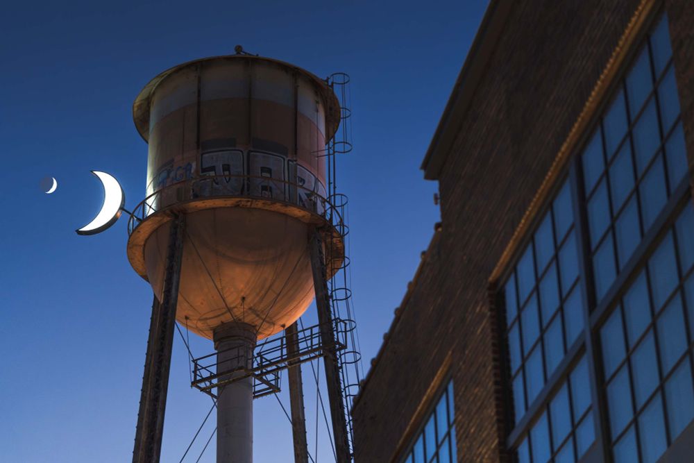 A water tower with a crescent moon light, juxtaposed next to the matching crescent moon in the sunset sky.