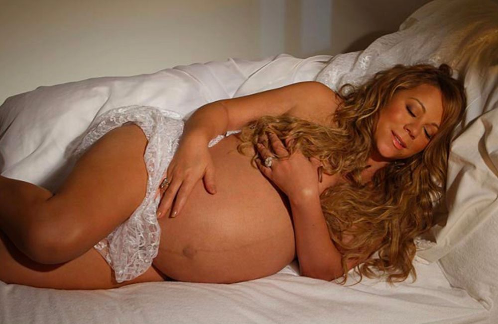 pregnant mariah carey laying on a bed holding her stomach while looking angelic