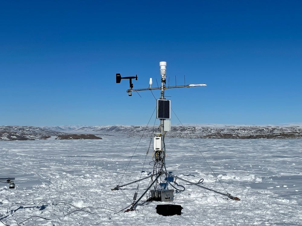 A weather station (KPC_L) standing on the Greenland Ice Sheet. The weather is sunny and mountains can be seen in the background