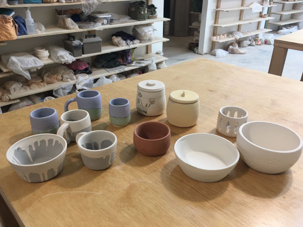 An assortment of glazed pottery on the studio table, shelving in the background. Some bowls, some mugs, some lidded jars, and a little votive holder. 