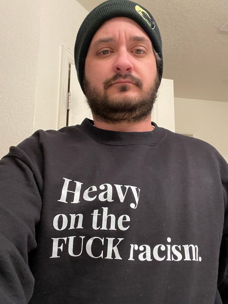 Adult male selfie wearing a black beanie hat and a black sweatshirt that says “Heavy on the FUCK racism.” 