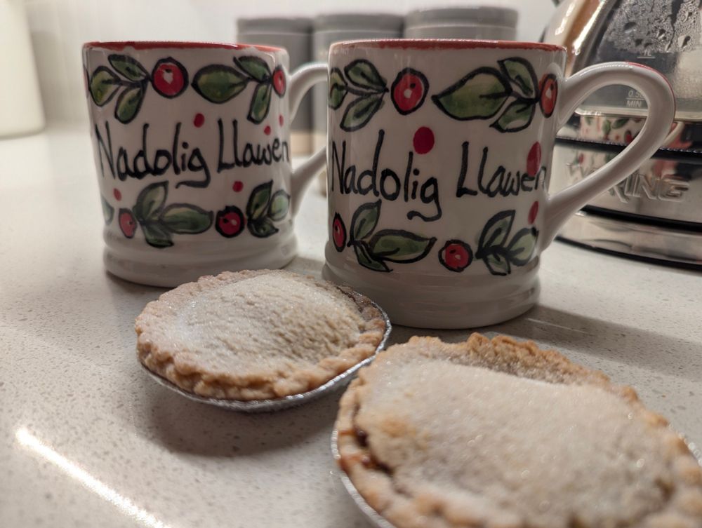 Two sugar-coated mince pies and two mugs with Nadolig Llawen written on them in a decorative style. 