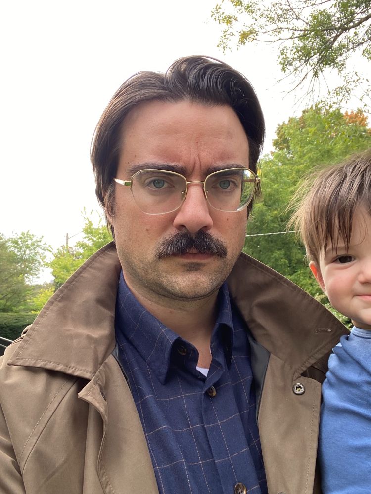 Outdoor selfie of a mustachioed man in a blue windowpane check button-up shirt and a trench coat carrying an absolutely adorable smiling baby on his arm. Overcast sky and trees and a power line are visible in the background. 