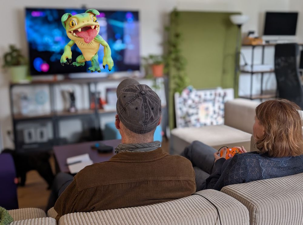 two people sitting on a couch with controllers, in front of a TV with blurred gameplay and a gremlin image slapped over it