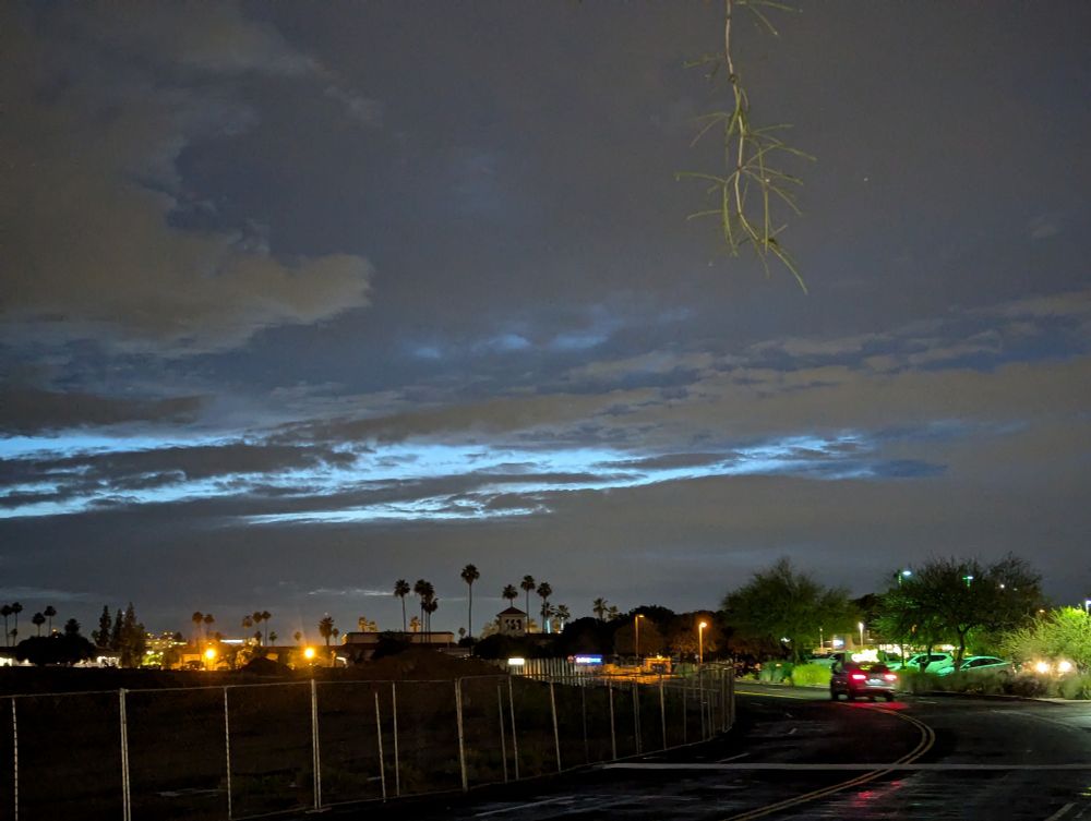 a picture of a dark, cloudy phoenix sky after rain. palm trees and lights from buildings are seen in the distance. a car drives along a road, illuminated and green.