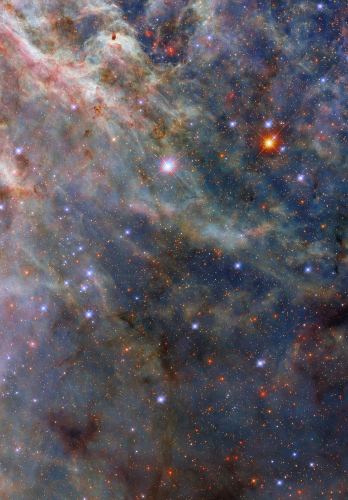 A nebula. The top-left is dense with layers of fluffy pink and greenish clouds. Long strands of green clouds stretch out from here; a faint layer of translucent blue dust combines with them to create a three-dimensional scene. A sparse network of dark dust clouds in the foreground adds reddish-black patches atop the nebula. Blue-white and orange stars, from our galaxy and beyond, are spread amongst the clouds.