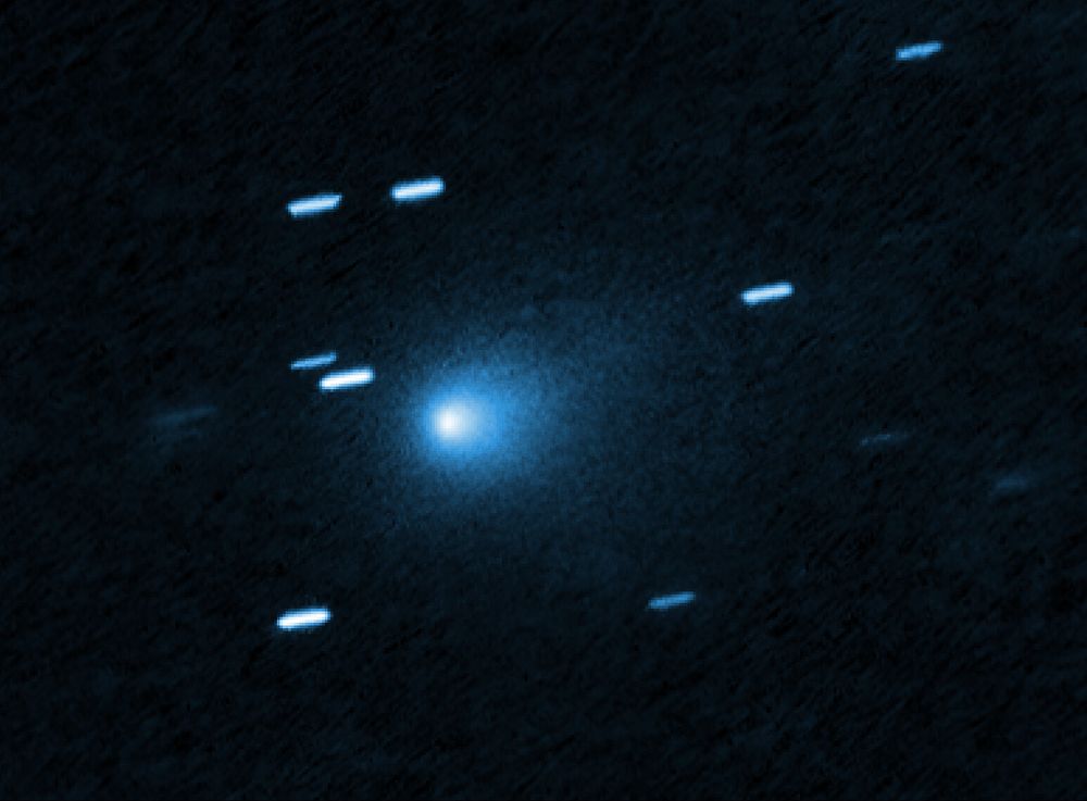 At the centre of the image is a comet that appears as a teardrop-shaped bluish cocoon of dust coming off the comet’s solid, icy nucleus and seen against a black background. The comet appears to be heading to the bottom left corner of the image. About a dozen short, light blue diagonal streaks are seen scattered across the image, which are from background stars that appeared to move during the exposure because the telescope was tracking the moving comet.