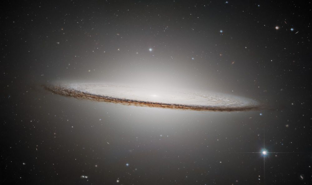 The Sombrero Galaxy is an oblong, pale white disc with a glowing core. It appears nearly edge-on but is slanted slightly in the front, presenting a slightly top-down view of the inner region of the galaxy and its bright core. The outer disc is darker with shades of brown and black. Different coloured distant galaxies and various stars are speckled among the black background of space surrounding the galaxy.
