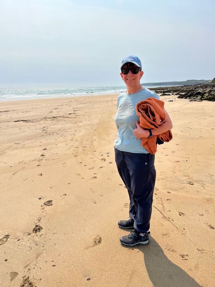Photo of me standing on sand, wearing hat, sunglasses and tee shirt, holding a rolled up orange towel 
