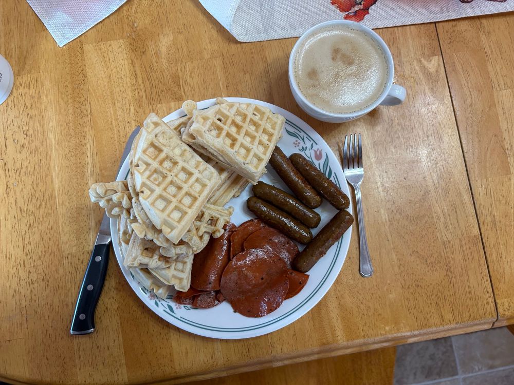 A white plate with a floral pattern on the edges, placed onto a wood table the plate has waffles, ham and sausage sitting next to the plate is a large mug containing americano 