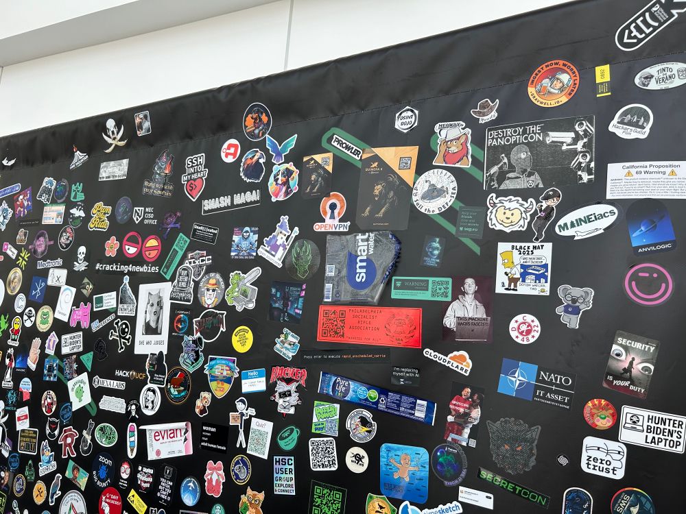 A big wall of stickers from a sticker wall at Def Con 32. In the upper middle right is a sticker of Jindall the Lion saying “Meowdy”