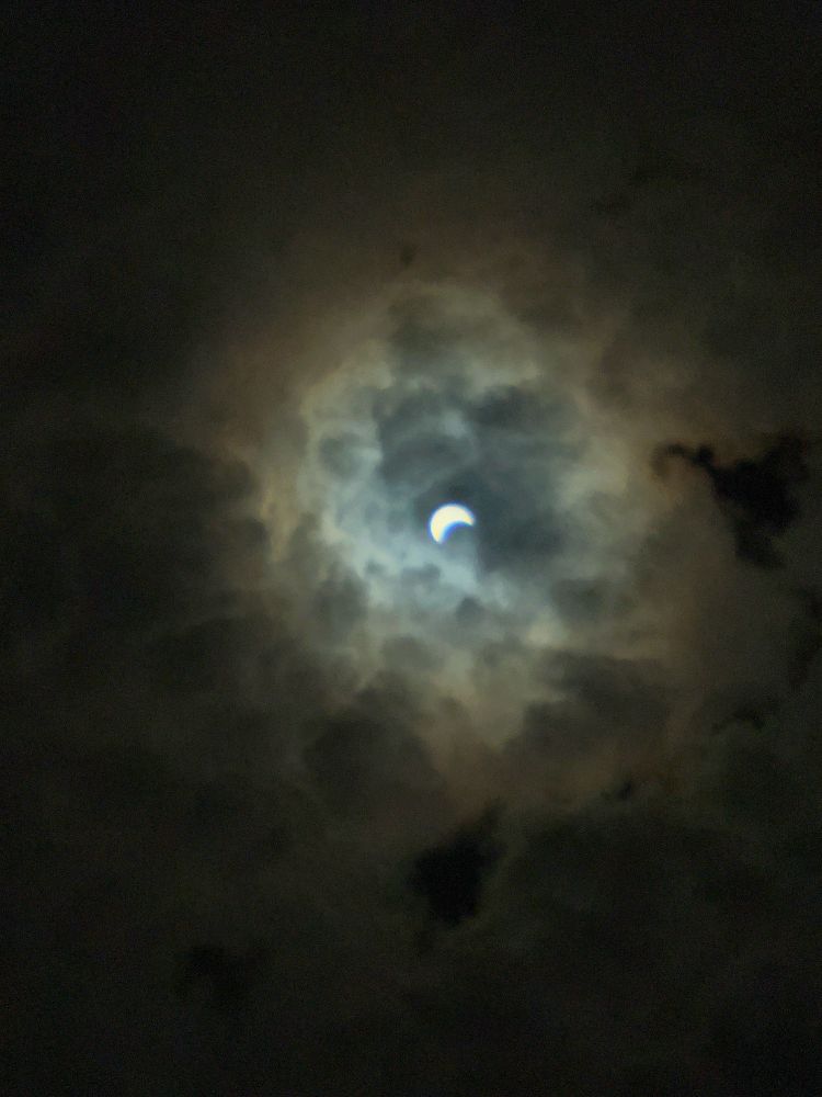 Photo of a crescent shape of the sun surrounded by glowing clouds. This is prior to the total eclipse.