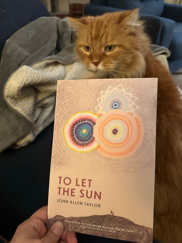 A photo of John Allen Taylor’s new collection of poetry “To Let The Sun” with my cat behind it.