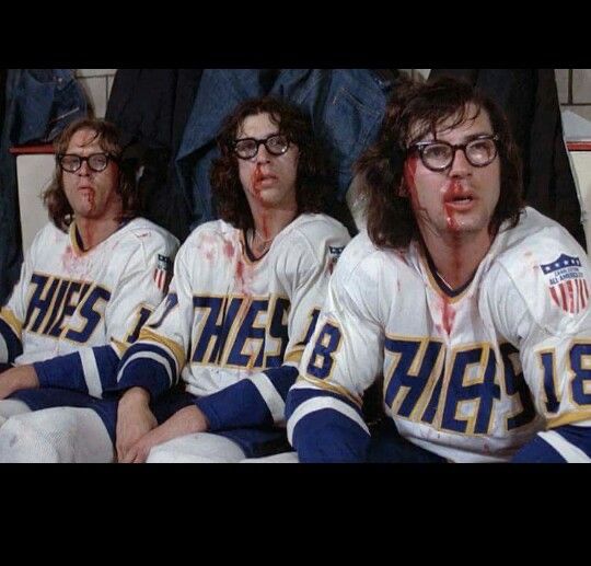 Three bloodied men with glasses, long hair, and hockey uniforms (the Charlestown Chiefs) looking to the right of the frame. They’ve been scrappin’!