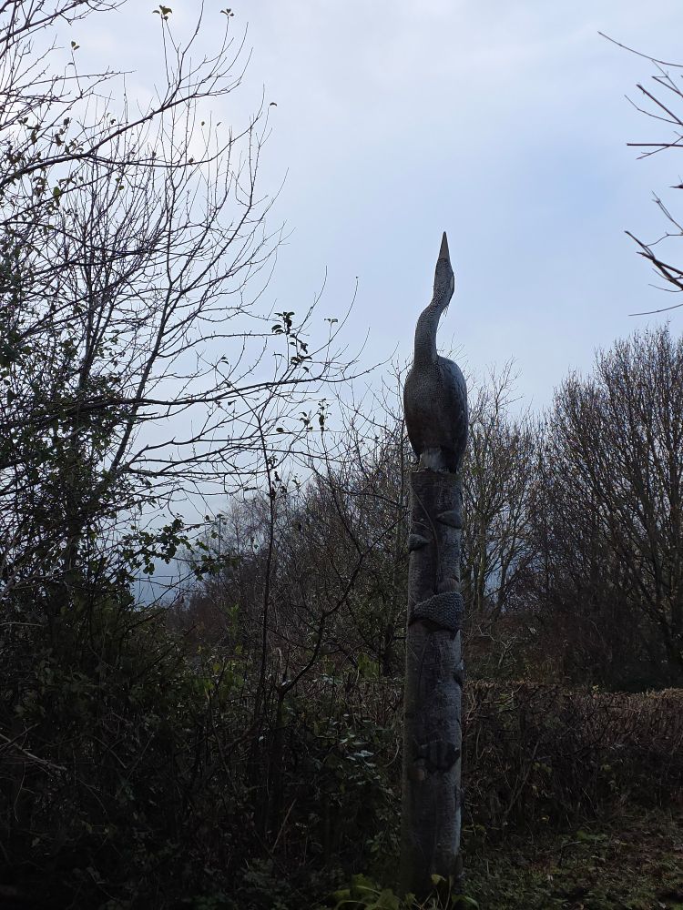 A wodden statue of a bird with a stiff neck surrpunded by bare trees and a moody sky.