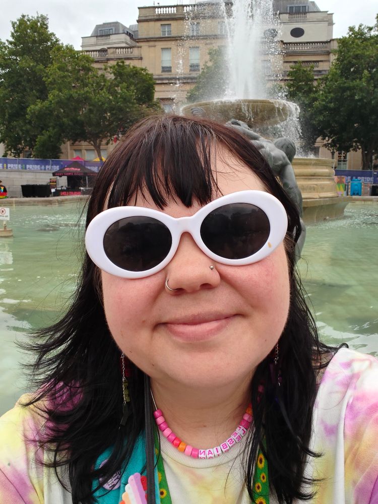 It's Rob sitting on a fountain in Trafalgar Square. They're wearing big white sunglasses and a necklace that reads "Lesbian", also a sunflower lanyard 