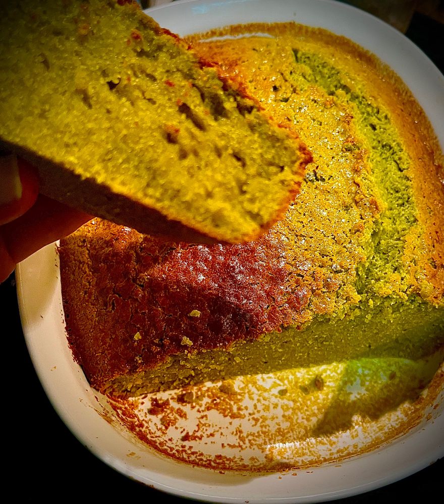 Massive slice of pale greenish cake w golden brown crust being held in foreground w rest of cake in white casserole in back