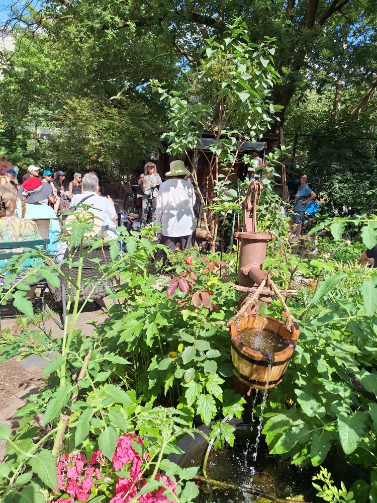 A fairy lush garden, some pink flowers and a fountain that looks like a water pump and a wooden bucket in the foreground, a klezmer band a bit in the distance.