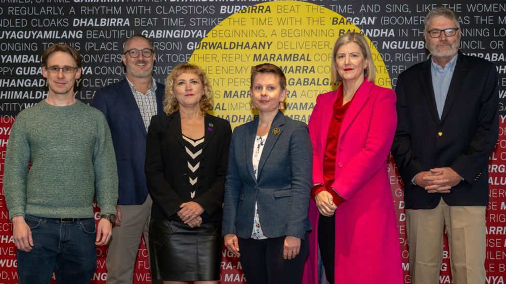 Daniel Nadasi (Google AU), David Liebowitz (PentenAmio), Emma Davidson (IDEATE Program Director), Penny Kyburz (ANU, IDEATE Steering Committee Chair), Sally-Ann Williams (Cicada Innovations, IDEATE Steering Committee member), Antony Hosking (ANU).
Standing in front of an Aboriginal flag that is overlaid with words in Ngunnawal and English.