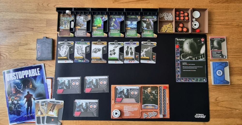 Top down shot of a card game set up on a table, including various decks of cards, tokens, rules, etc. 