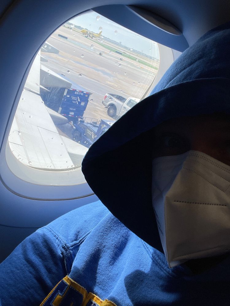 Me, in a blue hoodie and white mask sitting in an airplane window seat. Outside the window you can see the wing and some grounds crew