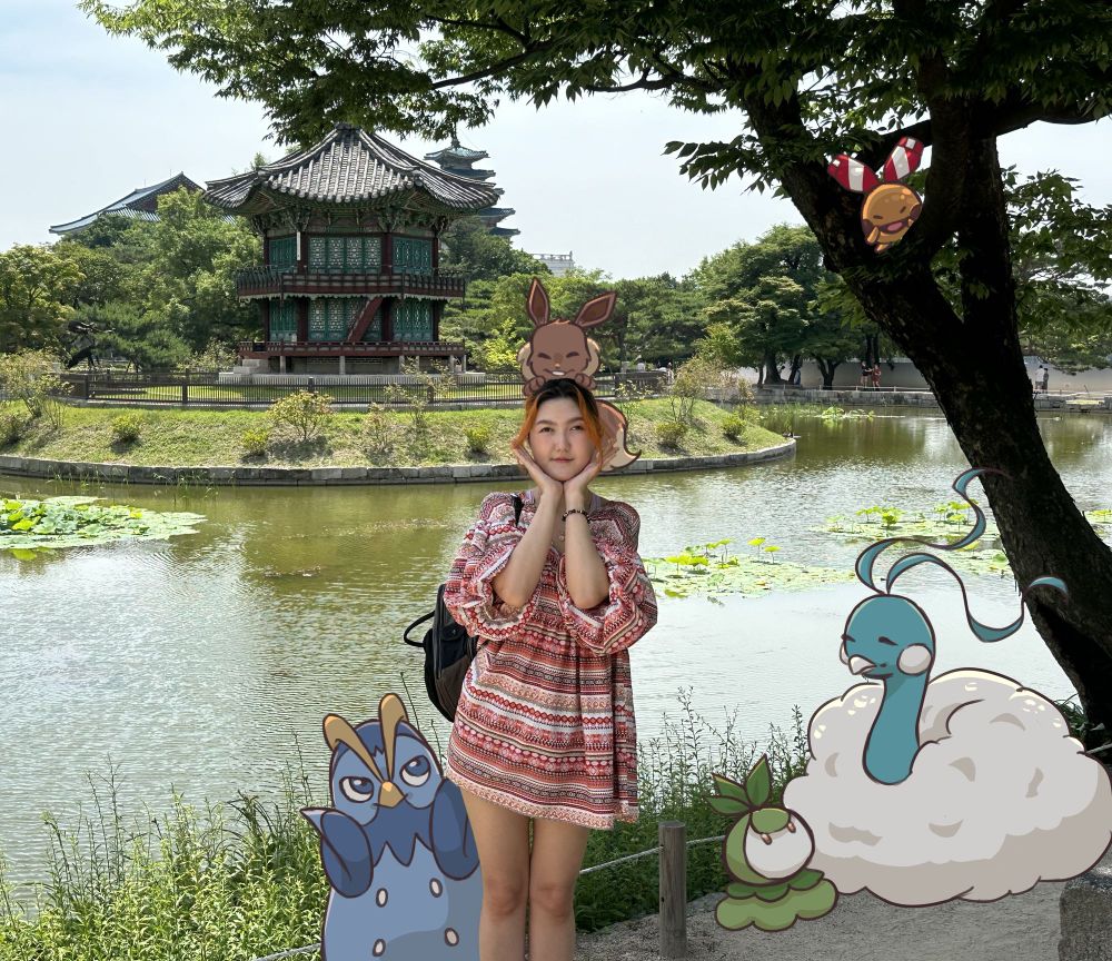 Young adult illustrated with prinplup mimicking face holding pose, eevee sitting on head, petilil sitting next to altaria, and chingling sitiing in tree. Historical korean palace buildings/lake in background.