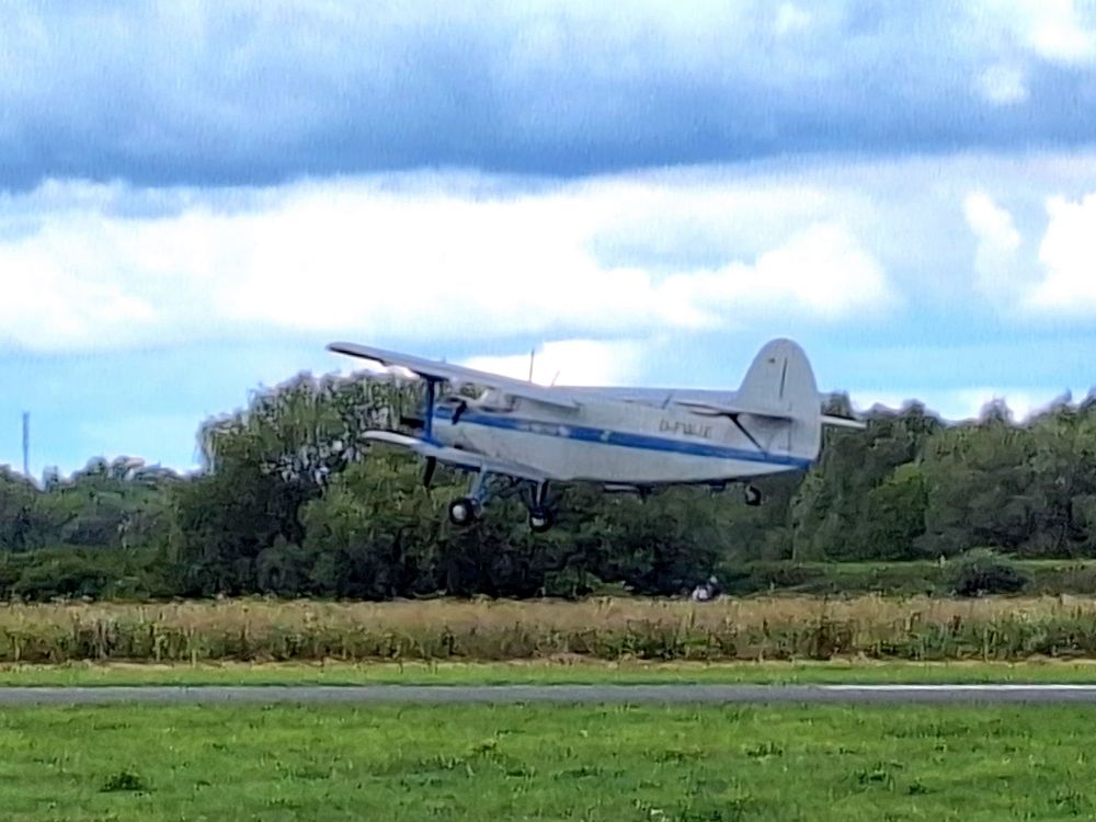 Man sieht auf einem kleinen Flugplatz einen wuchtigen silbernen Doppeldecker mit blauer 