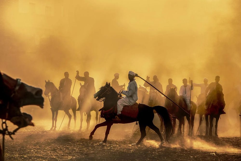 Photograph by Shams Saber Radwan of Egypt of a group of people on horseback, with their faces obscured by sunlight and dust; one is in the front of them and perpendicular to them. Photograph has warm yellow hue.