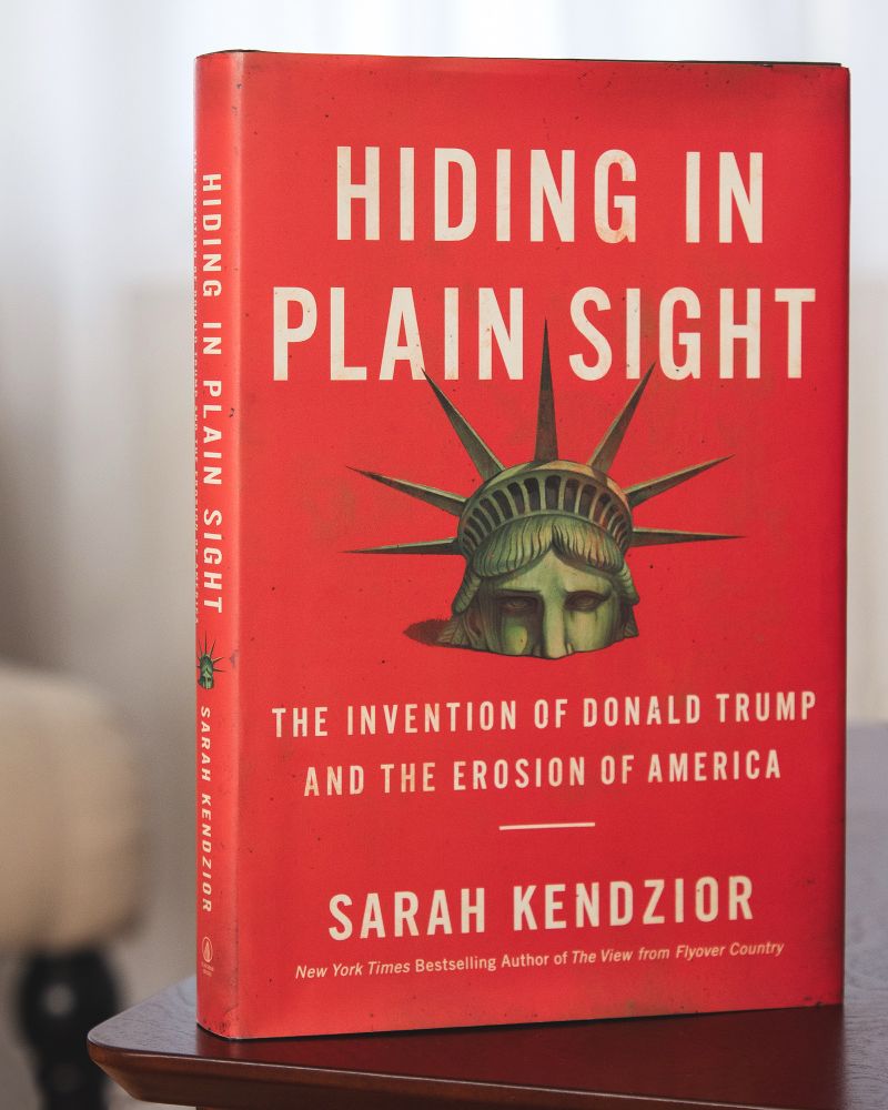 Hiding in Plain Sight by Sarah Kendzior. The book is standing on a mahogany end table. The book is reddish orange with title, subtitle and author name in white somewhat thin sans serif font. In the center of the book is the head of the Statue of Liberty; she appears to be suffocating and drowning.