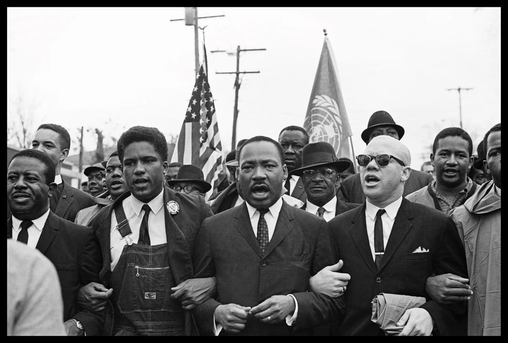 “(From left) Rev. Ralph Abernathy, James Forman, Dr. Martin Luther King Jr. and Rev. Jess Douglas lead the voting rights march to the Montgomery County Courthouse.”

Black and white photograph. Men’s arms are locked with flags in rear. James "Spider" Martin, 1965.