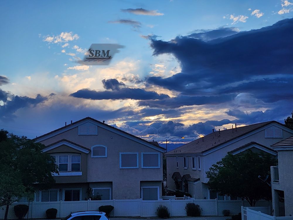 A complex design of cumulus, stratocumulus, and cumulonimbus clouds in colors of deep blue gray and illuminated yellow infront of a pale blue sky