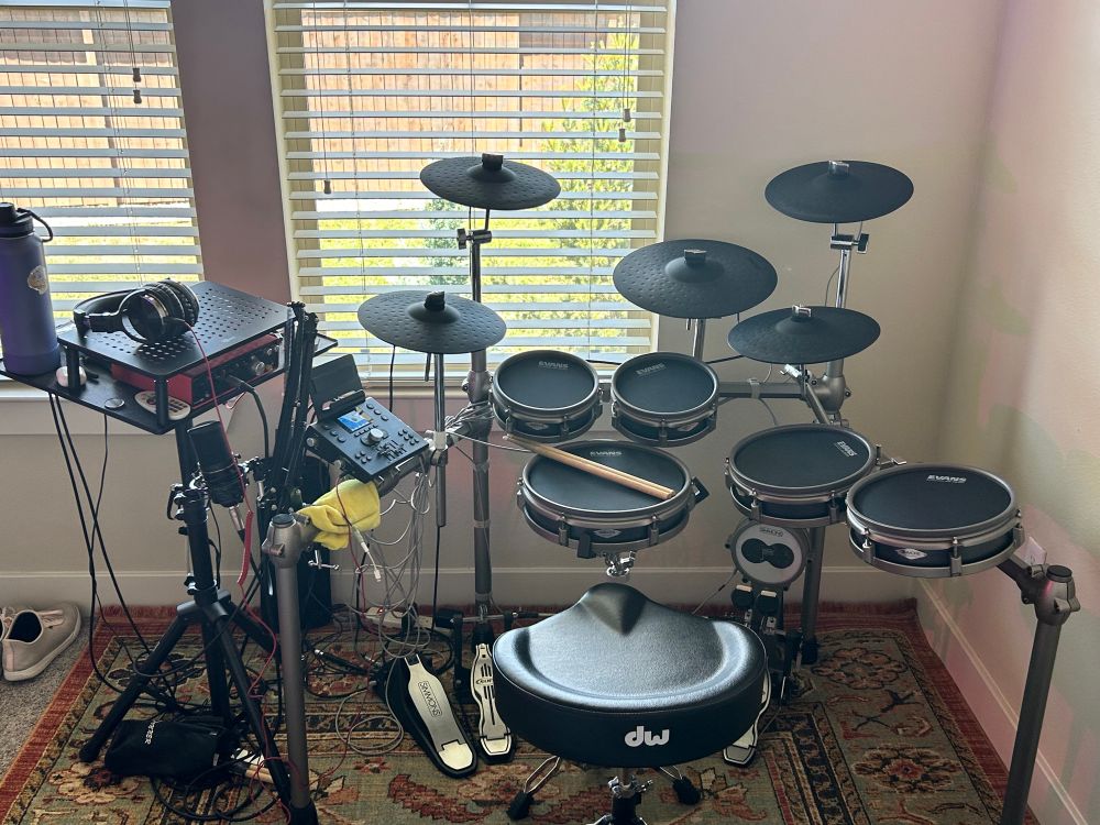 D_ROK’s electric drum kit with an extra hi-hat pad on the right side of the kit. There is a nest of cables and drum sticks on the floor.