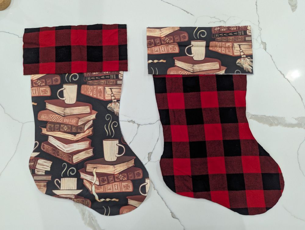 Two sets of fabric cut in the shape of Christmas stockings. One fabric has a black and red flannel pattern and the other features stacks of books with cups of tea on top.