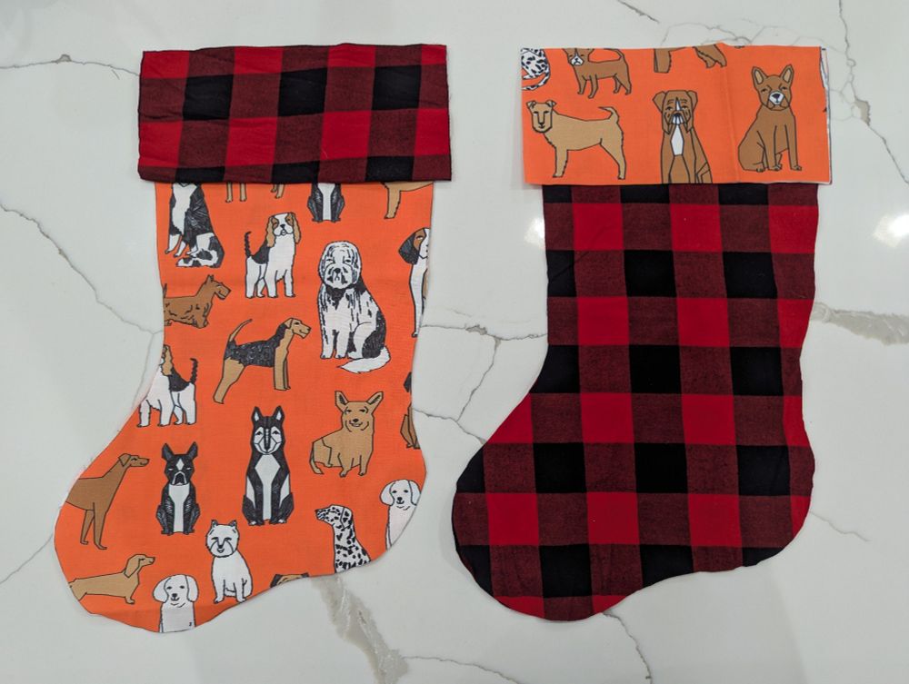 Two sets of fabric cut in the shape of Christmas stockings. One fabric has a black and red flannel pattern and the other features multiple dogs on an orange background.