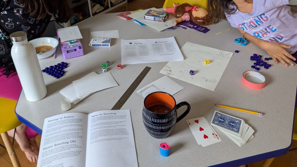 Table with supplies for playing The Deep Forest. Supplies included rules, playing cards, dice, and paper. Two children are seated at the table.
