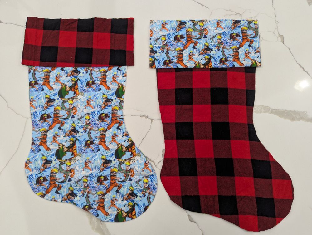 Two sets of fabric cut in the shape of Christmas stockings. One fabric has a black and red flannel pattern and the other features an anime character.