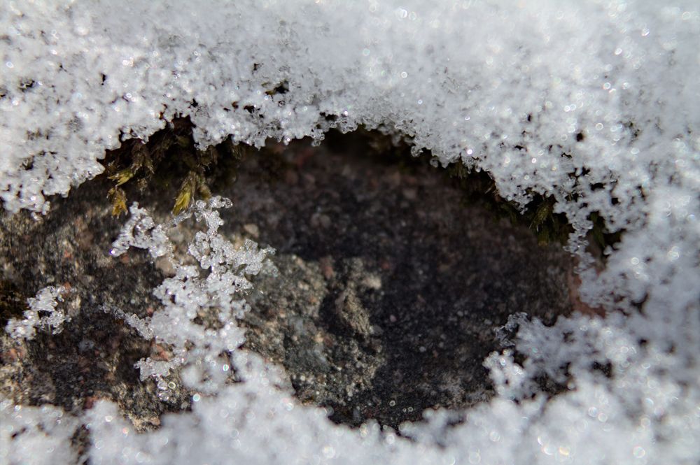 A patch of natural rock surrounded by snow crystals. There's a bit of moss poking from under the snow in the top-left part of the picture.