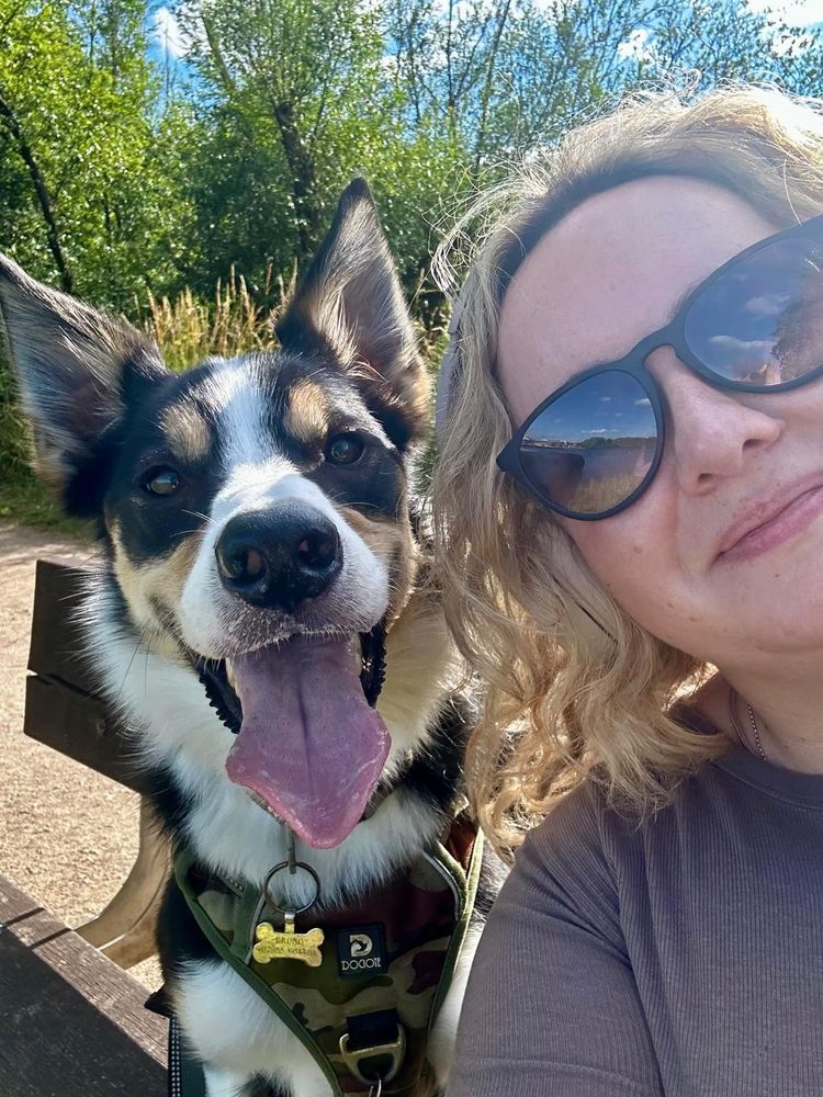 Abi (who has wavy blonde hair and sunglasses) smiling beside her happy dog, who has large, upright ears and is panting with his tongue out. The dog (Bruno) wears a camouflage harness. They are sitting on a bench outdoors on a sunny day, with greenery and blue sky in the background.