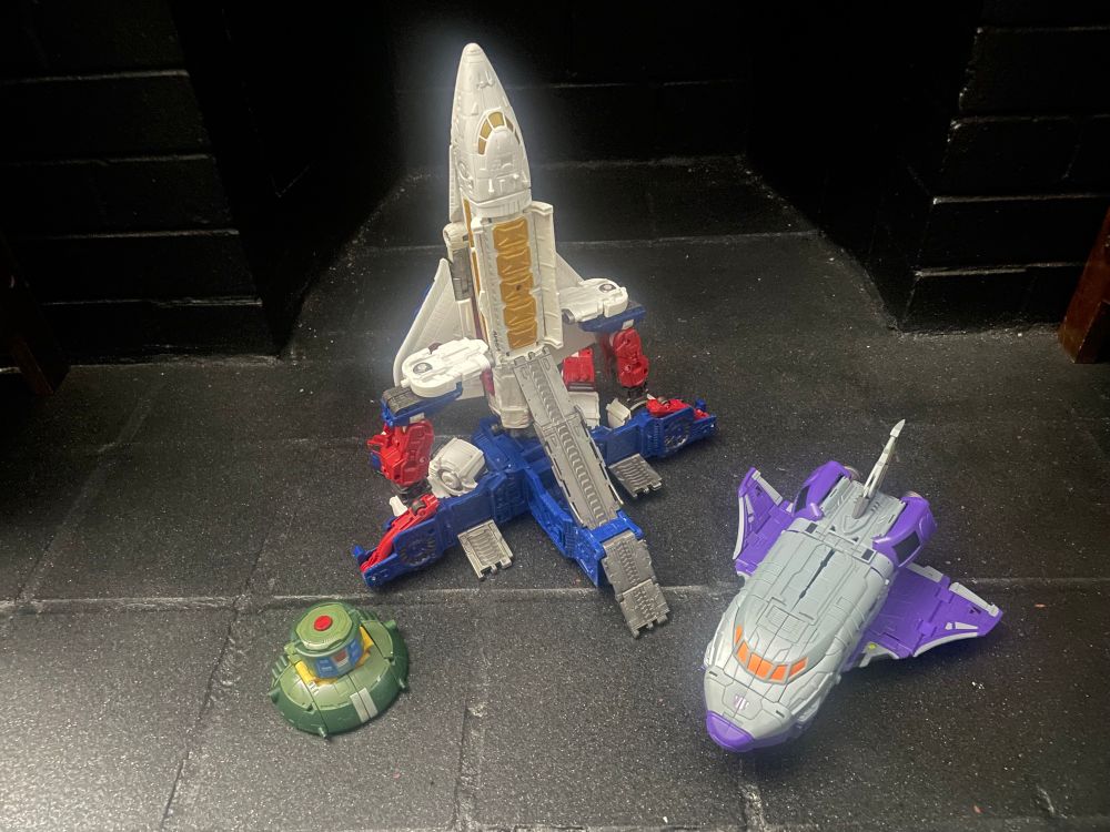 A bunch of space related toys, a ufo and two space shuttles with one on a launching pad 
Transformers legacy cosmos, earthrise Skylynx and dx9 chigurh (astrotrain)
