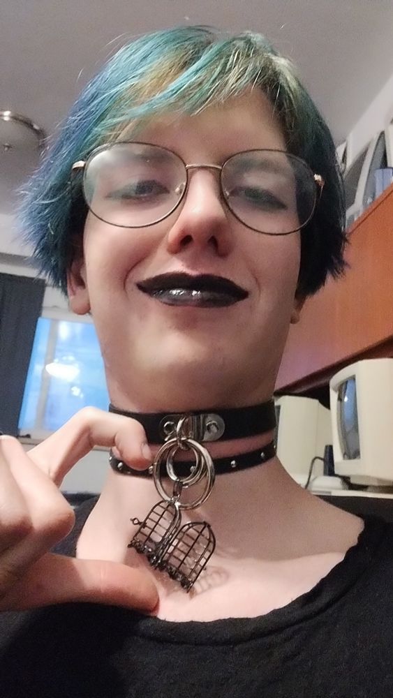 Selfie of Xae wearing two chokers, one of which has an open cage dangling from its rings. They are smirking and making half of a finger heart with their free hand next to the cage.